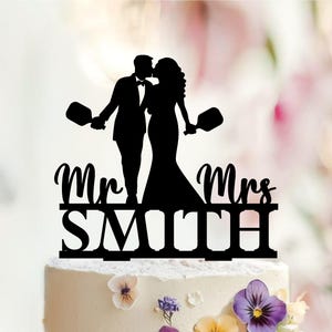 May include: A black silhouette cake topper featuring a couple in formal attire holding pickleball paddles. The text Mr Mrs SMITH is displayed in a combination of script and block lettering at the base of the figures, creating a decorative topper.