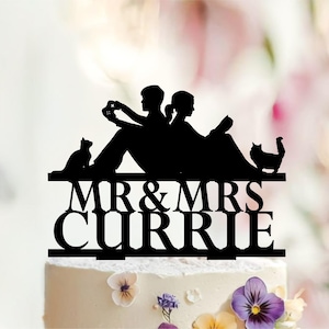 May include: A black cake topper silhouette featuring two people sitting back-to-back, one holding a game controller and the other a book, flanked by two cats. The text MR & MRS CURRIE is displayed in bold, capitalized letters below the figures.