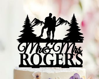 May include: A black cake topper featuring a silhouette of a couple standing in front of mountains and pine trees. Below the figures, the text reads Mr & Mrs ROGERS in a combination of cursive and block lettering, all mounted on a flat base.
