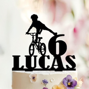 May include: A black cake topper featuring a silhouette of a person riding a bicycle with one leg extended, positioned next to the number 6 and the name LUCAS in bold capital letters, designed to be inserted into the top of a decorated cake.