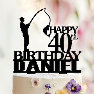 Może przedstawiać: Czarny topper na tort przedstawiający sylwetkę osoby łowiącej ryby z wędką i złowioną rybą. Napis brzmi HAPPY 40th BIRTHDAY DANIEL pogrubioną czarną czcionką. Dekoracja umieszczona jest na białym torcie ozdobionym kolorowymi kwiatami.