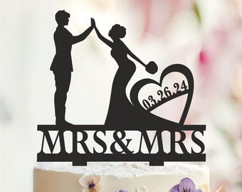 May include: Black silhouette cake topper featuring two figures, a bride and a groom, high-fiving. The design includes a heart with the date 03.26.24 and the text "MRS & MRS" below. The topper is placed on a decorated cake.