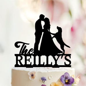 Pode incluir: Um topo de bolo em silhueta preta apresentando um casal frente a frente com um cão a saltar ao lado. O texto "The REILLY'S" está integrado na base do design. O topo está posicionado sobre um bolo com cobertura branca.