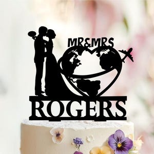 May include: A black cake topper featuring a silhouette of a couple facing each other, a heart-shaped globe with an airplane, the text MR&MRS, and the name ROGERS at the base. The design is flat and mounted on a white cake decorated with flowers.