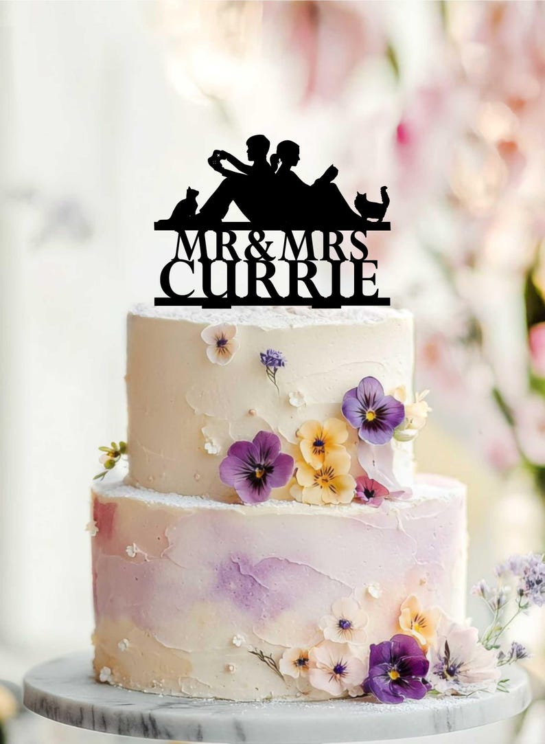 May include: A black silhouette cake topper featuring two people sitting and reading books with two cats, positioned above the text MR & MRS CURRIE. The topper is placed on a two-tier cake decorated with purple, yellow, and white pansy flowers.