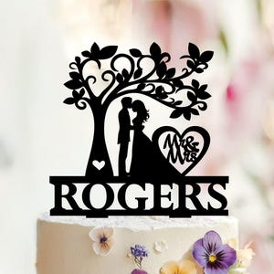 Peut inclure: Un gâteau topper noir représentant la silhouette d'un couple sous un arbre. Un cœur contenant le texte Mr & Mrs est placé à côté du couple. La base de la décoration affiche le nom ROGERS en lettres capitales grasses et bien visibles.