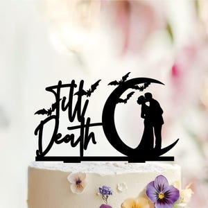 May include: A black cake topper featuring the text 'Till Death' in a script font, accompanied by silhouettes of several flying bats and a couple kissing while standing on a crescent moon. The design is mounted on two flat base prongs.