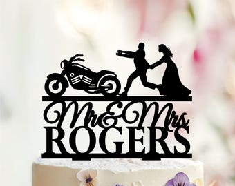 May include: A black cake topper featuring a silhouette of a motorcycle, a person running, and another person following. Below the figures, the text reads Mr & Mrs ROGERS in a combination of cursive and block lettering, all set on a flat base.