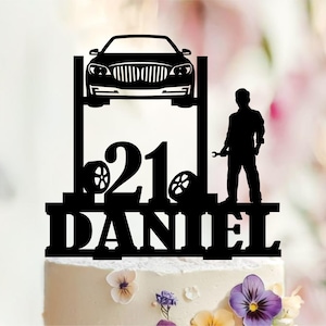 May include: A black cake topper featuring a silhouette of a car on a lift, the number 21, two tyres, a mechanic holding a spanner, and the name DANIEL in bold capital letters. The design is a flat, laser-cut style piece intended for cake decoration.