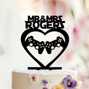 May include: A black cake topper featuring the text MR & MRS ROGERS in a stylized font above a heart outline. Inside the heart, two black video game controllers are positioned facing each other, creating a symmetrical design for a wedding cake.