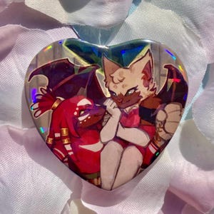 May include: A heart-shaped pin featuring a holographic finish and an illustration of two anthropomorphic characters. One character is red with spiky hair, while the other is cream-coloured with dark wings. The design includes vibrant colours and sharp lines.
