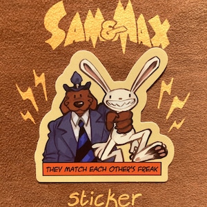 Peut inclure: Un autocollant découpé représentant les personnages Sam et Max. Un chien marron en costume bleu et cravate tient un lapin blanc au large sourire. Le texte Sam & Max figure au-dessus, et le texte THEY MATCH EACH OTHER'S FREAK est dans un encadré orange.