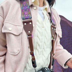 May include: A brown leather camera strap with a colourful woven pattern. The strap is attached to a black DSLR camera with a lens.