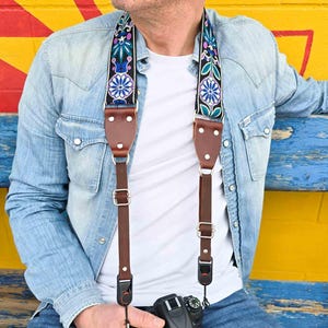 May include: A brown leather camera strap with a colourful floral pattern. The strap is adjustable and has a quick-release buckle. The strap is being worn by a person wearing a blue denim shirt.