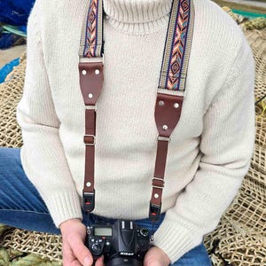 May include: A brown leather camera strap with a colourful woven pattern. The strap is attached to a camera and worn by a person.