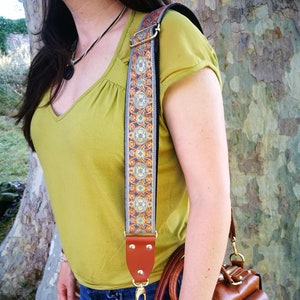 May include: A brown leather handbag with a decorative strap featuring a geometric pattern in orange, blue, and gold. The bag has a top handle and a zipper closure.