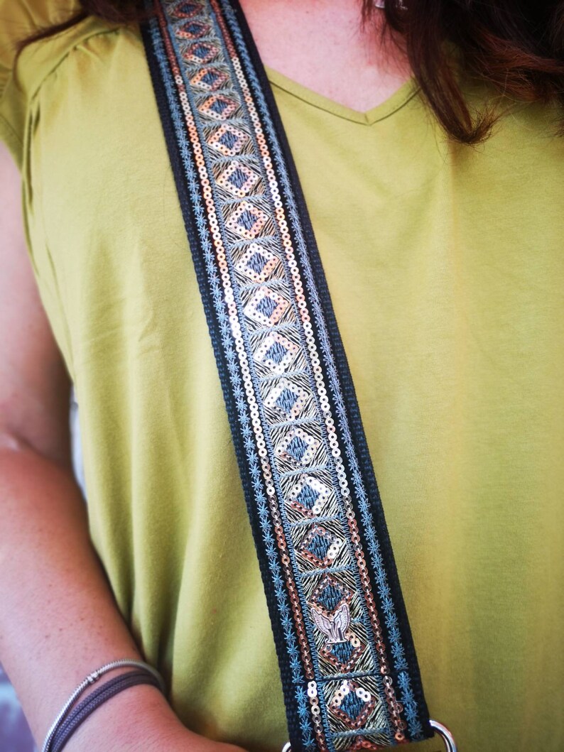 May include: A black guitar strap with a blue and gold geometric pattern. The strap is decorated with sequins and has a silver buckle.