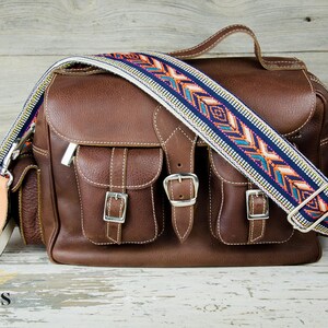 Pode incluir: Uma bolsa de c&acirc;mera de couro marrom com dois bolsos frontais e uma al&ccedil;a estampada. A bolsa tem fivelas prateadas e um z&iacute;per prateado.