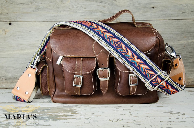 Pode incluir: Uma bolsa de c&acirc;mera de couro marrom com dois bolsos frontais e uma al&ccedil;a estampada. A bolsa tem fivelas prateadas e um z&iacute;per prateado.