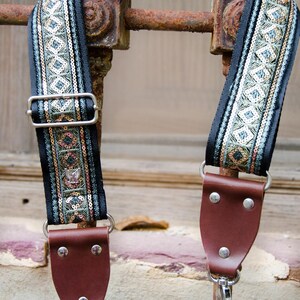 May include: A black camera strap with a green, gold, and silver patterned trim. The strap has brown leather ends with metal hooks.