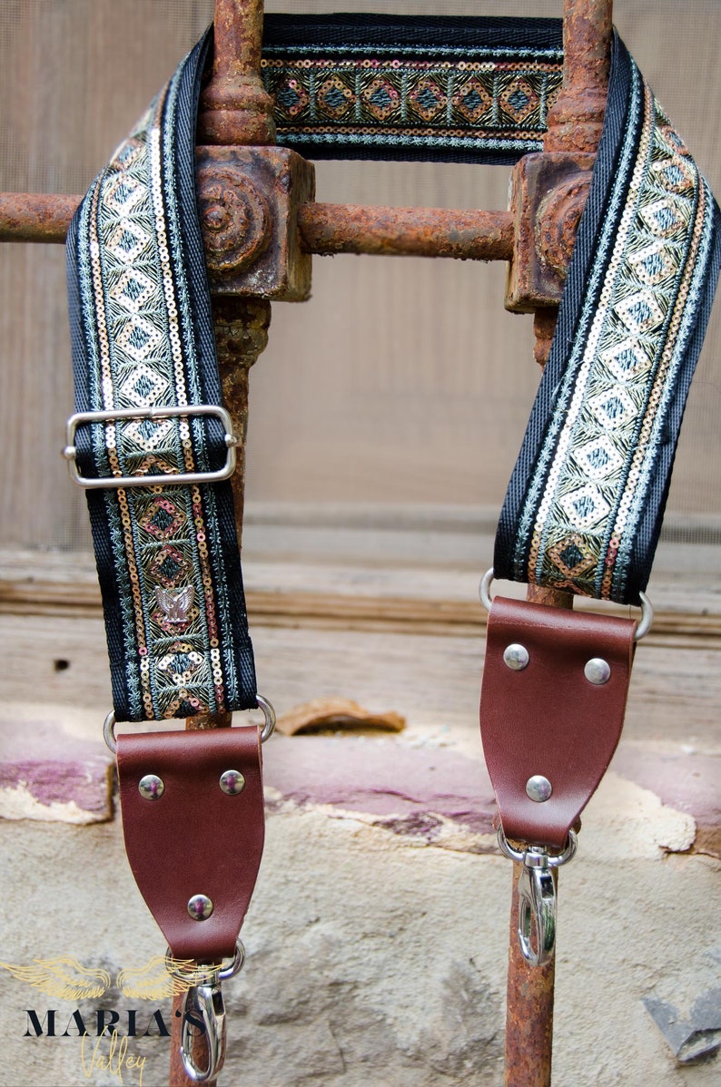 May include: A black camera strap with a green, gold, and silver patterned trim. The strap has brown leather ends with metal hooks.