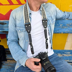 May include: A black and white patterned camera strap with leather accents. The strap is worn by a person holding a black camera with a silver lens. The camera has the word "Nikon" printed on it.