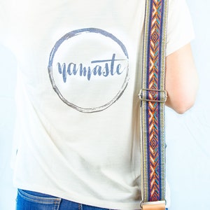 Pode incluir: Uma camiseta branca com um gr&aacute;fico circular que diz "namaste" em uma fonte cinza. A camiseta est&aacute; sendo usada por uma pessoa com uma al&ccedil;a estampada colorida sobre o ombro.