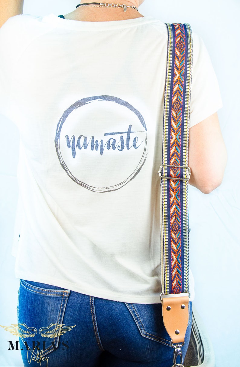 Pode incluir: Uma camiseta branca com um gr&aacute;fico circular que diz "namaste" em uma fonte cinza. A camiseta est&aacute; sendo usada por uma pessoa com uma al&ccedil;a estampada colorida sobre o ombro.