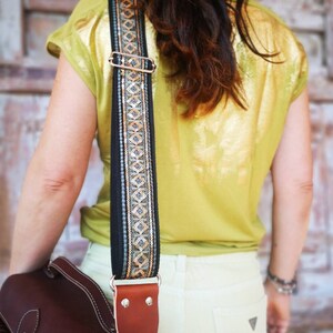May include: A brown leather camera bag with a black and gold patterned strap. The strap is adjustable and has a metal buckle. The bag is worn over the shoulder of a person wearing a yellow shirt and white pants.