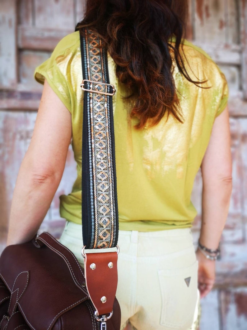 May include: A brown leather camera bag with a black and gold patterned strap. The strap is adjustable and has a metal buckle. The bag is worn over the shoulder of a person wearing a yellow shirt and white pants.