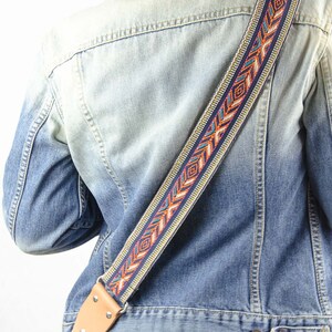 Pode incluir: Uma pessoa vestindo uma jaqueta de jeans azul com uma correia de guitarra estampada colorida. A correia tem uma ponta de couro marrom com uma fivela de metal.