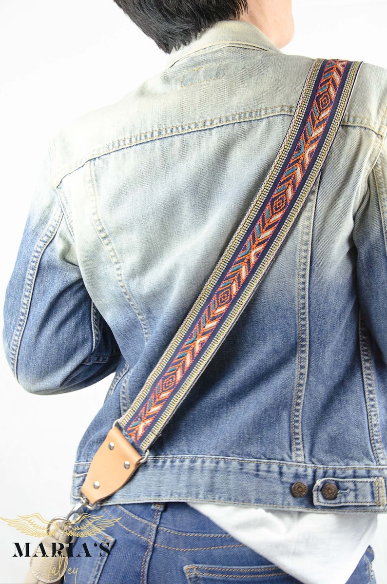 Pode incluir: Uma pessoa vestindo uma jaqueta de jeans azul com uma correia de guitarra estampada colorida. A correia tem uma ponta de couro marrom com uma fivela de metal.