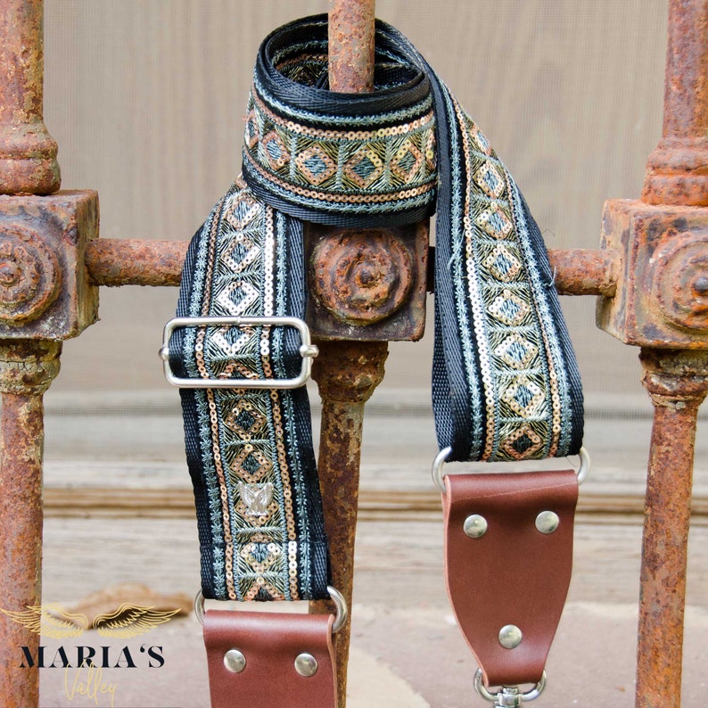 May include: A black camera strap with a green and gold geometric pattern. The strap has silver metal hardware and brown leather ends.