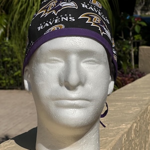 May include: A black scrub cap featuring the Baltimore Ravens logo in purple and gold. The cap has a purple band and ties in the back. The cap is displayed on a white mannequin head.
