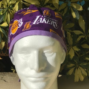 May include: A purple scrub cap featuring a repeating Los Angeles Lakers basketball team logo and text pattern. The cap has a solid light purple fabric band along the bottom edge and is displayed on a white mannequin head for clear visibility.