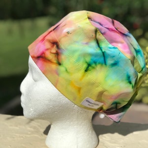 May include: A surgical cap featuring a vibrant tie-dye pattern with blended hues of yellow, pink, blue, and green. The cap includes a small white rectangular fabric label on the side with the text GOSIASURGICALCAPS and Etsy.com printed in black.
