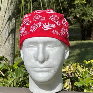 May include: A red cap with the word "Indiana" in white script repeated across the fabric. The cap is displayed on a white mannequin head, showcasing the design and fit. The cap has a red trim.