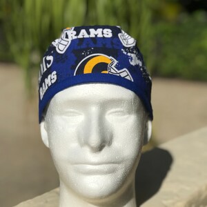 May include: A scrub cap featuring a dark blue background with a repeating pattern of the word RAMS in white block letters and yellow and blue football helmet graphics. The cap has a solid blue fabric band along the bottom edge.