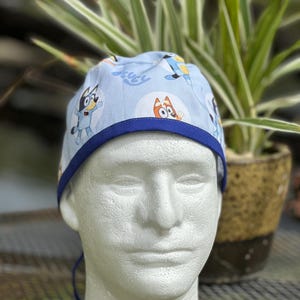 May include: Blue and white surgical scrub hat with a cartoon dog print. The hat has a blue trim and a tie string.