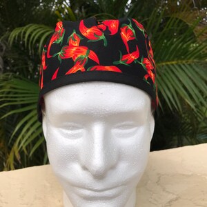 May include: A black scrub cap featuring a vibrant pattern of red chili peppers with green stems. The cap has a solid black fabric band along the bottom edge and a fitted crown design, providing full coverage for the head with a clean, finished look.
