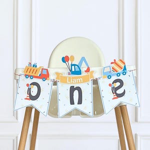 May include: A high chair banner featuring three pennants with the letters O, N, and E in dark gray. The banner displays construction vehicle illustrations, colorful balloons, and a yellow ribbon banner with the name Liam printed in white text.