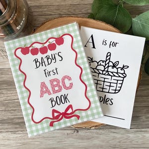 May include: A Baby's First ABC Book with a light green gingham background, red apple illustrations, and a red bow. Beside it is a white page with black text reading A is for and an illustration of a basket of apples with the word ples visible.