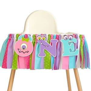 May include: A colorful high chair banner with the word "ONE" in purple, blue, and green letters. The banner is decorated with three monster faces, one pink, one purple, and one blue.