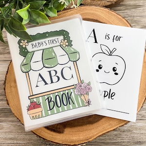 May include: A children's ABC book with a cover featuring a green and white awning design with the text "BABY'S FIRST ABC BOOK." The book is open to a page with an apple illustration and the text "A is for apple."