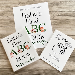 Puede incluir: Tres libros blancos para beb&eacute;s con el texto "Baby's First ABC Book" y "A is for acorn". Los libros tienen ilustraciones de un zorro, setas y hojas. El fondo es una superficie de madera.