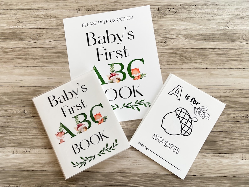 Puede incluir: Tres libros blancos para beb&eacute;s con el texto "Baby's First ABC Book" y "A is for acorn". Los libros tienen ilustraciones de un zorro, setas y hojas. El fondo es una superficie de madera.