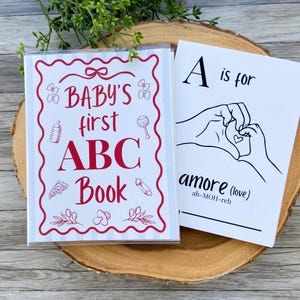 May include: A white book cover with red text reading BABY'S first ABC Book, surrounded by red line drawings of baby items. Beside it, a white page with black text reading A is for amore (love) and a line drawing of hands forming a heart.