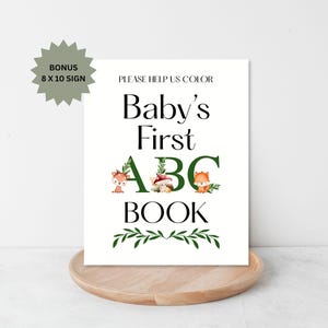 Puede incluir: Un letrero blanco con el texto "Baby's First ABC Book" en negro y verde. El letrero incluye ilustraciones de animales del bosque y vegetaci&oacute;n. Se incluye un letrero adicional de 20,3 x 25,4 cm.