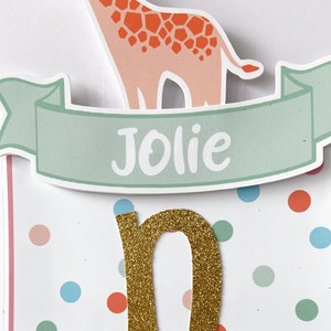 May include: A banner with a giraffe illustration and the name "Jolie" in white on a light green banner. The letter "n" is gold glitter. The background is white with colorful polka dots.
