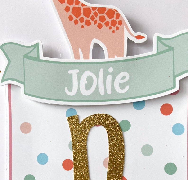 May include: A banner with a giraffe illustration and the name "Jolie" in white on a light green banner. The letter "n" is gold glitter. The background is white with colorful polka dots.